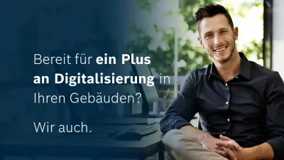 Ein lächelnder Mann in dunklem Hemd sitzt an einem Schreibtisch vor einem Computerbildschirm. Im Bild der Text: 'Bereit für ein Plus an Digitalisierung in Ihren Gebäuden? Wir auch.'