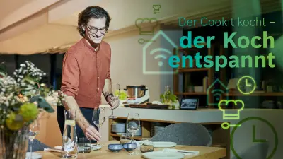 Martin Strumpler deckt einen Esstisch mit Gläsern und Besteck. Im Hintergrund kocht der Cookit das Risotto.