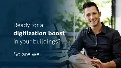 A smiling man in a dark shirt sits at a desk in front of a computer screen. In the image the text reads: 'Ready for digitalization boost in your buildings? So are we.