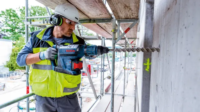 Bauarbeiter benutzt eine Bohrmaschine an einer Betonwand, während er auf einem Gerüst steht.