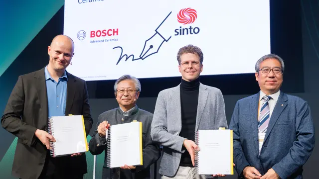 Four people holding signed documents in front of Bosch Advanced Ceramics and Sinto logos.
