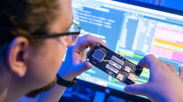 Person holding a small electronic circuit board in front of a computer screen.