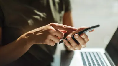 Nahaufnahme der Hände einer Person, die ein Smartphone hält und damit interagiert, im Hintergrund ist ein Laptop zu sehen.