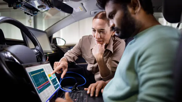 Two people are sitting inside a car where they are looking at a laptop and testing their developments.