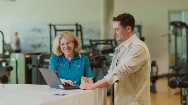 Zwei Personen arbeiten gemeinsam an einem Laptop im Empfangsbereich eines Fitnessstudios.