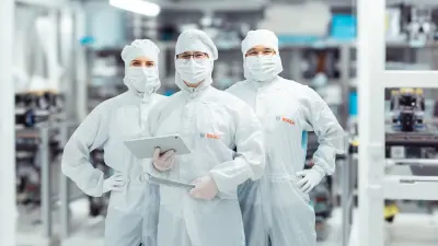 Drei Personen in weißen Schutzanzügen in einer Produktionshalle.