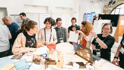 Eine Gruppe steht lachend um einen Tisch und arbeitet gemeinsam an verschiedenen Aufgaben beim Hackathon „Bosch Connected Experience“ in Berlin. Der Tisch ist vollgepackt mit bunten Notizzetteln, Stiften und einem Laptop. Im Hintergrund stehen weitere Teilnehmer um Pinnwände, an denen verschiedene Projekt Plakate hängen.