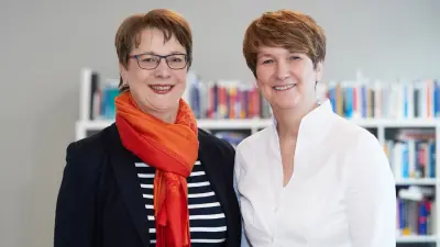Zwei Frauen stehen vor einem Bücherregal und lächeln in die Kamera.