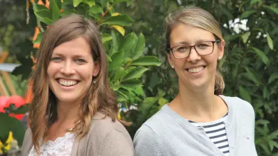 Zwei Frauen stehen vor Pflanzen und lächeln in die Kamera.