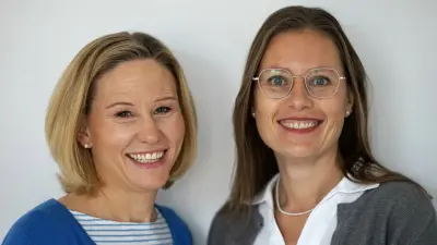 Zwei Frauen stehen vor einer weißen Wand und lächeln in die Kamera.
