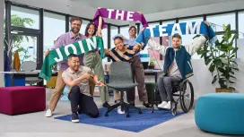 A diverse group is holding scarves with the inscription “Join the Team” in the air and is pointing to an empty office chair.