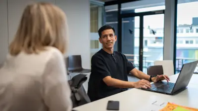 Antonio smiles and wears a dark shirt. He is working on his laptop and conversing with a female colleague in the office.