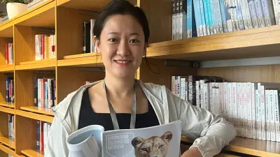 Mengfei smiles and holds the "Harvard Business Review" in a library with wooden bookshelves filled with books.