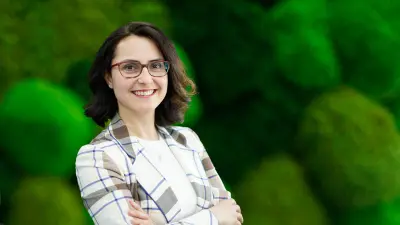 Nagihan trägt einen karierten Blazer und hat ihre Arme verschränkt. Sie steht vor einer grünen Mooswand.