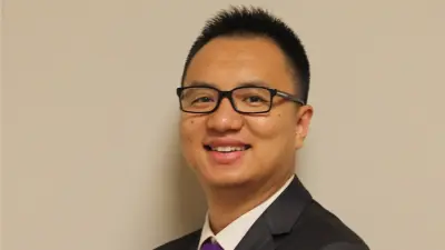 Ruobing is smiling, wearing a suit and a purple tie, with glasses, and standing against a light background.