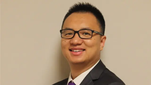 Ruobing is smiling, wearing a suit and a purple tie, with glasses, and standing against a light background.