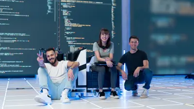 A team of students is sitting on the floor laughing. One student has a laptop on her lap, another shows a peace sign. The third sits in a squatting position. Code can be seen in the background.