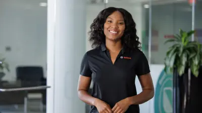 A woman stands in an office and looks into the camera with a smile.