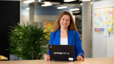 Aline Deloir sitzt, gekleidet in einen blauen Blazer an einem Tisch mit ihrem Laptop, auf dem ein #LikeABosch-Sticker klebt. Im Hintergrund sieht man eine Pflanze, einen Terminplaner und eine Pinnwand.