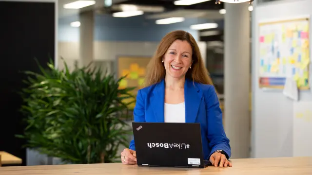 Aline Deloir sitzt, gekleidet in einen blauen Blazer an einem Tisch mit ihrem Laptop, auf dem ein #LikeABosch-Sticker klebt. Im Hintergrund sieht man eine Pflanze, einen Terminplaner und eine Pinnwand.