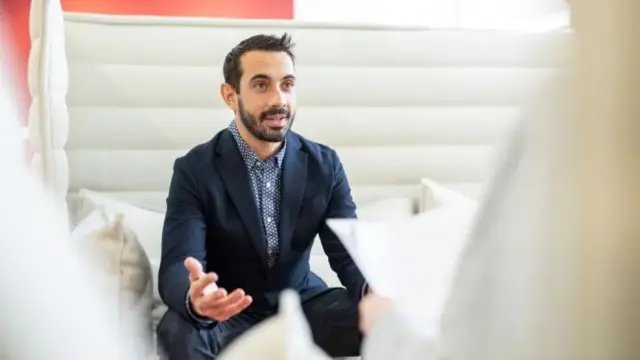 André Luis Tournoux, sitting in a seating group on a white sofa. He is wearing a suit and talking gesticulating to his counterpart, who cannot be seen.