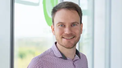 Andreas Frischen trägt ein gemustertes Hemd, steht vor einer Glasscheibe und lächelt in die Kamera.