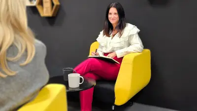 Angela sits in a yellow chair with a notebook and pen, leading a conversation. Two coffee mugs are on the table.