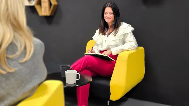 Angela sits in a yellow chair with a notebook and pen, leading a conversation. Two coffee mugs are on the table.