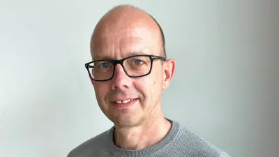 Christian is wearing a gray shirt and black glasses. He is standing in front of a white wall, looking into the camera and smiling slightly.