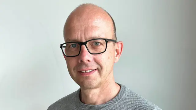 Christian is wearing a gray shirt and black glasses. He is standing in front of a white wall, looking into the camera and smiling slightly.