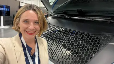 Christine is wearing a cream-colored blazer, smiles in the camera takes a selfie in front of a vehicle.