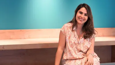 Damla has brown hair and is wearing a summery dress. She sits smiling on a wooden bench in front of a turquoise wall.