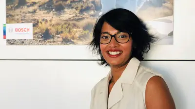 Estelle Donet-Selvan steht vor einer Wand, an der ein Bosch-Poster hängt. Sie trägt eine hellbeige Bluse und eine Brille und lächelt in die Kamera.