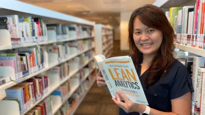 Gan Bee Keen standing between two bookshelves, holding a book titled "Lean Analytics" and smiling at the camera.