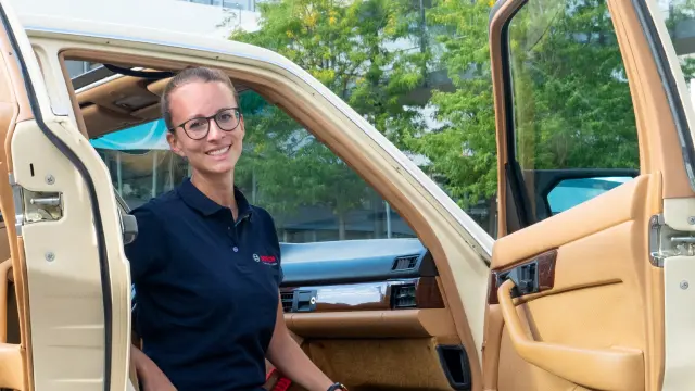 Hanna Späth sitzt in einem beigen Auto, die Türen sind geöffnet, und blickt nach draußen zur Kamera. Sie trägt ein blaues Bosch-Poloshirt und eine Brille.