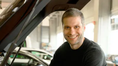 Holger Banzhaf befindet sich neben einem aufgeklappten Kofferraum eines schwarzen Autos und lächelt in die Kamera. Im Hintergrund ist ein weiteres Auto sichtbar in der Farbe weiß.