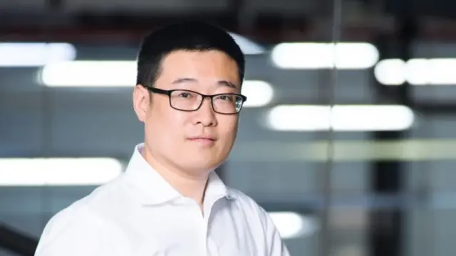 Portrait of Honghui Wu wearing a white blouse and black glasses. In the background you can see various reflections of lamps.