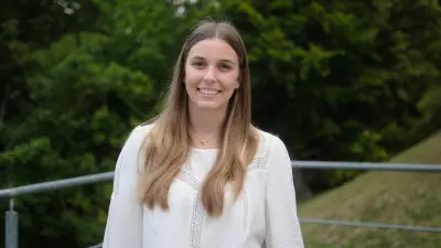 Jana Kremer trägt eine weiße Bluse und steht draußen, lächelnd, vor einem Geländer. Dahinter sieht man ein Feld und viele Bäume.