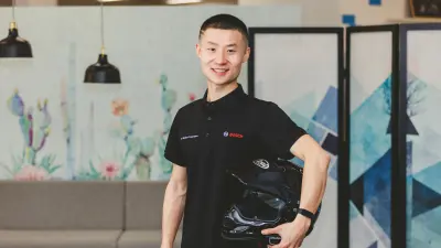 Jerry Gan wears a black Bosch polo shirt, holds a motorbike helmet and smiles at the camera. A modern office can be seen in the background.