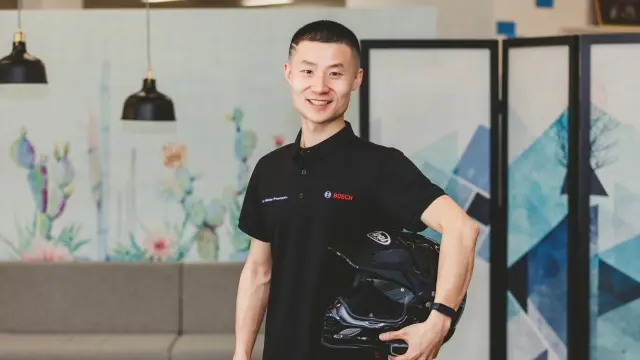 Jerry Gan wears a black Bosch polo shirt, holds a motorbike helmet and smiles at the camera. A modern office can be seen in the background.
