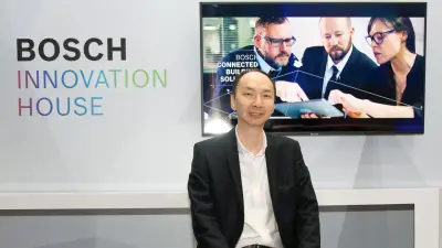 Joseph Hong sitzt vor einer Wand, an der ein Bildschirm befestigt ist, der Menschen zeigt, und die Aufschrift „Bosch Innovation House" zu lesen ist. Er trägt Hemd und Anzug und lächelt in die Kamera.
