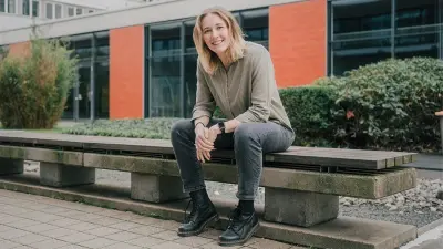 Katharina Fleischer-Dörr can be seen in an outdoor area. She sits on a long bench and looks into the camera with a smile. In the background, you can see a building with orange walls and large windows, and green bushes in front of it. 