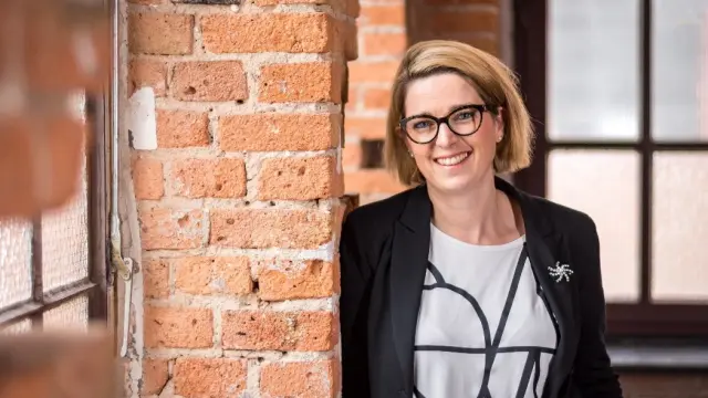 Katharina Hopp lehnt an einer Säule aus Ziegelsteinen. Sie trägt ein groß-gemustertes weißes T-Shirt, einen Blazer und eine Brille und lächelt in die Kamera.