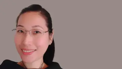 Mai Lan Ha wears glasses and a ponytail, stands in front of a wall and smiles at the camera.
