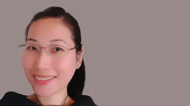 Mai Lan Ha wears glasses and a ponytail, stands in front of a wall and smiles at the camera.