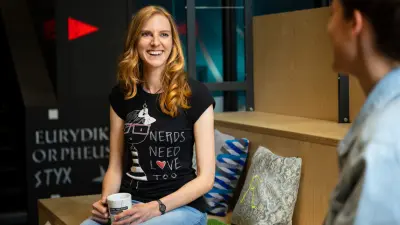 Marita Klein sitzt mit einer Person gegenüber von ihr auf einer Holzbank mit mehreren bunten Kissen. Sie trägt eine Jeans und ein schwarzes T-Shirt und hält eine Tasse in der Hand. Sie lächelt ihr Gegenüber an.