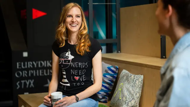 Marita Klein sitzt mit einer Person gegenüber von ihr auf einer Holzbank mit mehreren bunten Kissen. Sie trägt eine Jeans und ein schwarzes T-Shirt und hält eine Tasse in der Hand. Sie lächelt ihr Gegenüber an.
