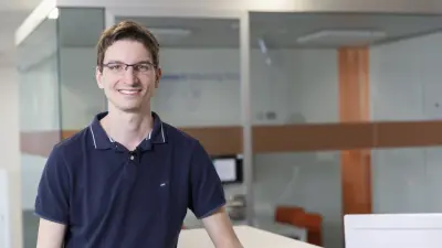 Michael Schäfer steht an einen weißen Stehtisch gelehnt und lacht in die Kamera. Er trägt ein blaues Polo-Shirt, eine Armbanduhr und eine Brille.