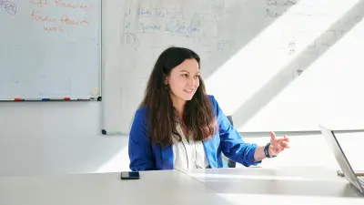 Patricia is sitting at a table in an office, with whiteboards written on in the background.