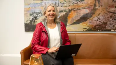 Petra Kama-Welle sitzt auf einem hellbraunen Ledersofa und trägt eine rote Lederjacke über einem weißen T-Shirt. Im Hintergrund sieht man ein orange-schwarz-blaues Bild.